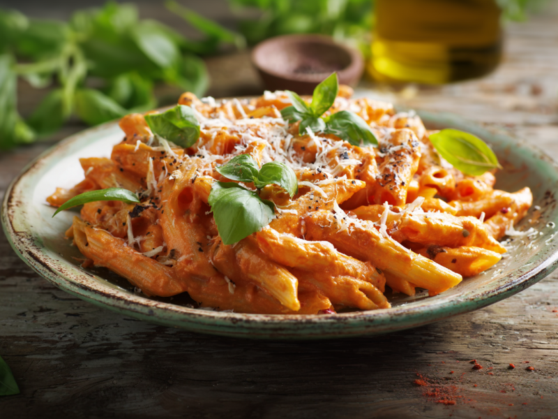 Tomaten-Ricotta-Nudeln auf einem rustikalen Teller auf einem Holztisch. Rundum den Teller und im Hintergrund sieht man frische Basilikumblätter liegen.