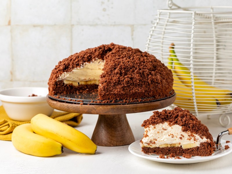 Ein veganer Maulwurfkuchen auf einem Präsentierteller. Ein Stück ist herausgeschnitten und gibt den Blick auf die Füllung frei. Davor und dahinter liegen insgesamt vier Bananen.