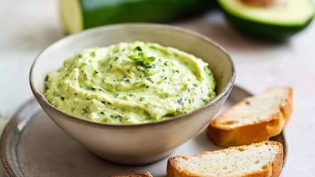 Schale mit Zucchini-Avocado-Aufstrich, Scheiben Brot daneben und halbe Avocado und eine Zucchini im Hitergrund