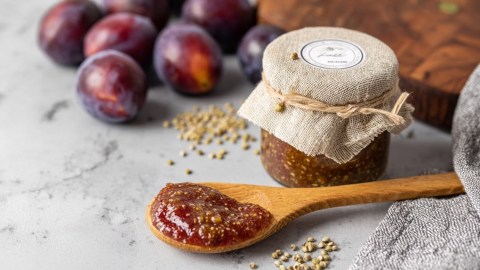 Ein kleines Einmachglas mit Zwetschgen-Senf. Davor liegt ein Holzlöffel mit Zwetschgen-Senf. Im Hintergrund sind einige frische Zwetschgen zu sehen.