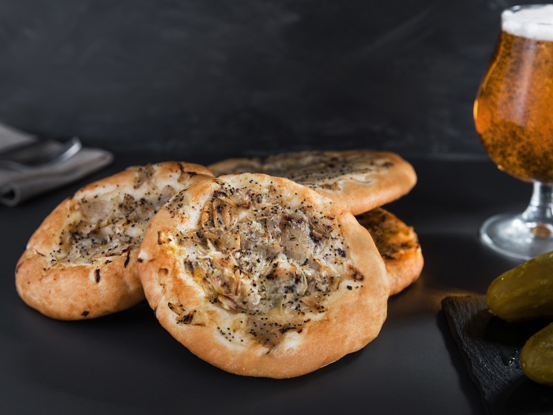 Vier Cebularz angerichtet auf einem Tisch. Rechts neben ihnen steht ein Bier im Glas. Davor liegen einige Gewürzgurken.