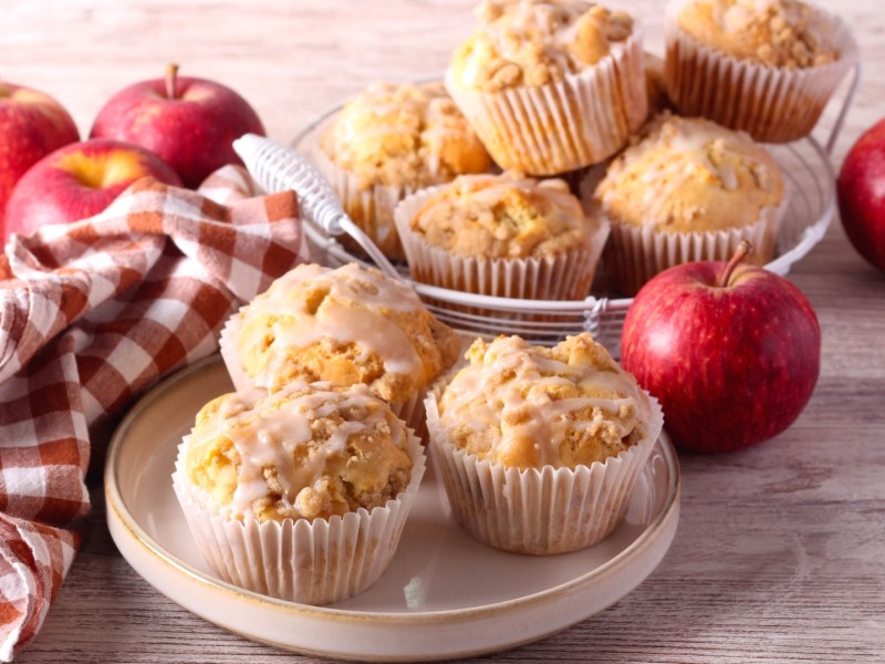 Mehrere Apfelmuffins mit Sour Cream auf zwei Tellern. Daneben liegen frische Äpfel.