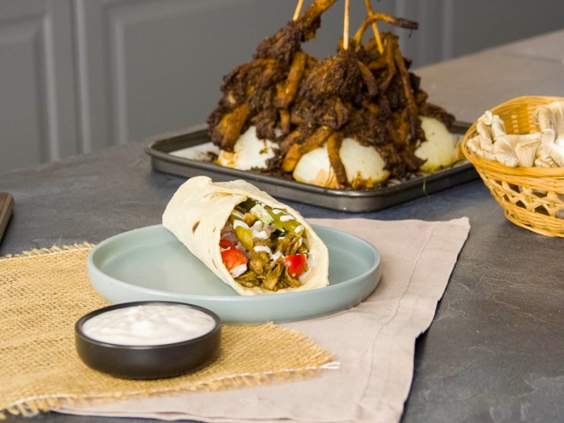 Ein Wrap, gefüllt mit Austernpilz-Schawarma. Im Hintergrund sieht man die restlichen Schawarma-Spieße. Vor dem Wrap steht eine Schale mit Joghurtsoße.