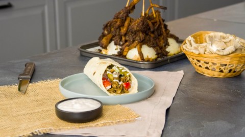 Ein Wrap, gefüllt mit Austernpilz-Schawarma. Im Hintergrund sieht man die restlichen Schawarma-Spieße. Vor dem Wrap steht eine Schale mit Joghurtsoße.