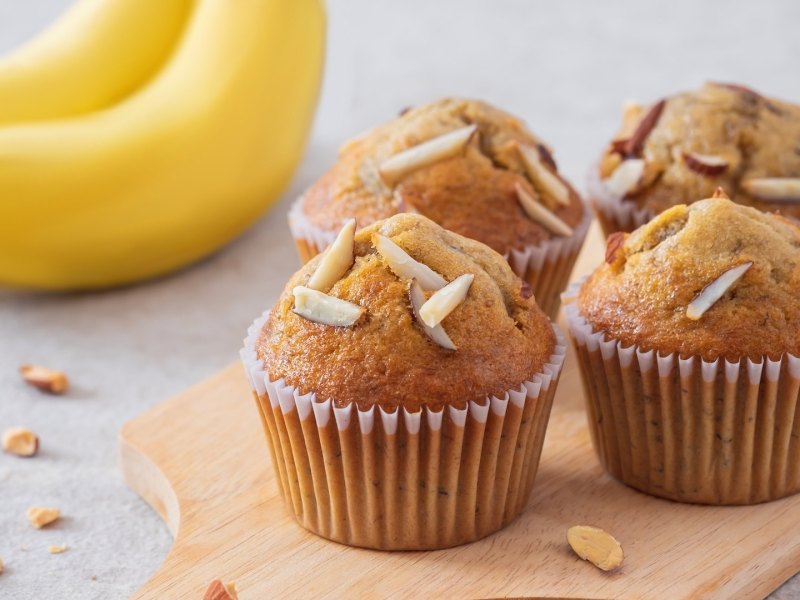 Mehrere Bananenmuffins aus dem Airfryer auf einem Holzbrett, daneben zwei Bananen.