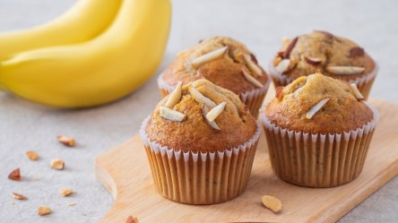 Mehrere Bananenmuffins aus dem Airfryer auf einem Holzbrett, daneben zwei Bananen.