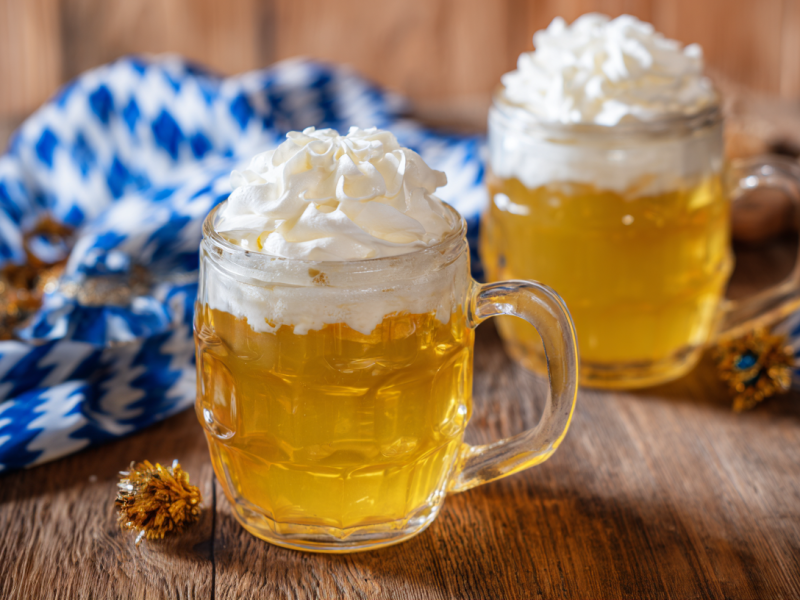 Zwei Gläser, die mit Bier-Wackelpudding gefüllt sind auf einem Holztisch. Neben den Gläsern liegen kleine, goldene Dekobällchen und traditionelle bayerische Platzdeckchen.