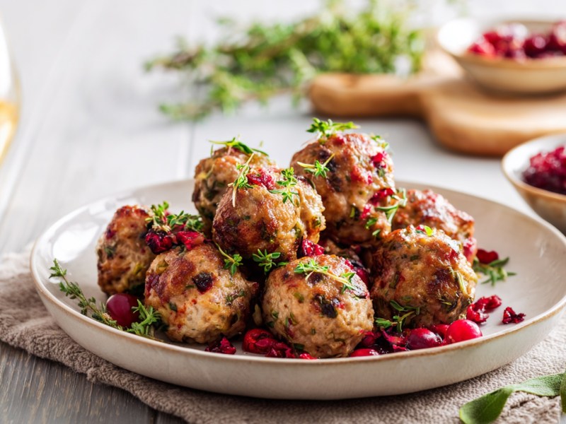 Ein Teller mit Fleischbällchen mit Kräuter und Cranberrys. Im Hintergrund kann man ein Glas Weißwein sowie Schalen mit Cranberrys und Cranberrysoße und Kräutern erkennen.