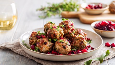 Ein Teller mit Fleischbällchen mit Kräuter und Cranberrys. Im Hintergrund kann man ein Glas Weißwein sowie Schalen mit Cranberrys und Cranberrysoße und Kräutern erkennen.