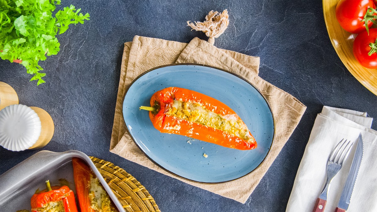 Ein blauer Teller mit einer gefüllten Paprika mit Feta und Kräutern. Ringsum kann man eine Stoffserviette mit Besteckt, frische Kräuter, Paprika und einen Salzstreuer erkennen.