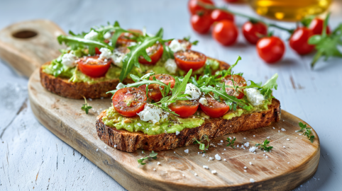 Zwei Scheiben Gourmet-Toast auf einem kleinen Holzbrett angerichtet. Sie sind mit Avocado, Ziegenkäse, Tomaten und Rucola belegt. Man sieht deutlich das grobe Salz, das über sie gestreut wurde. Im Hintergrund liegen weitere Tomaten,