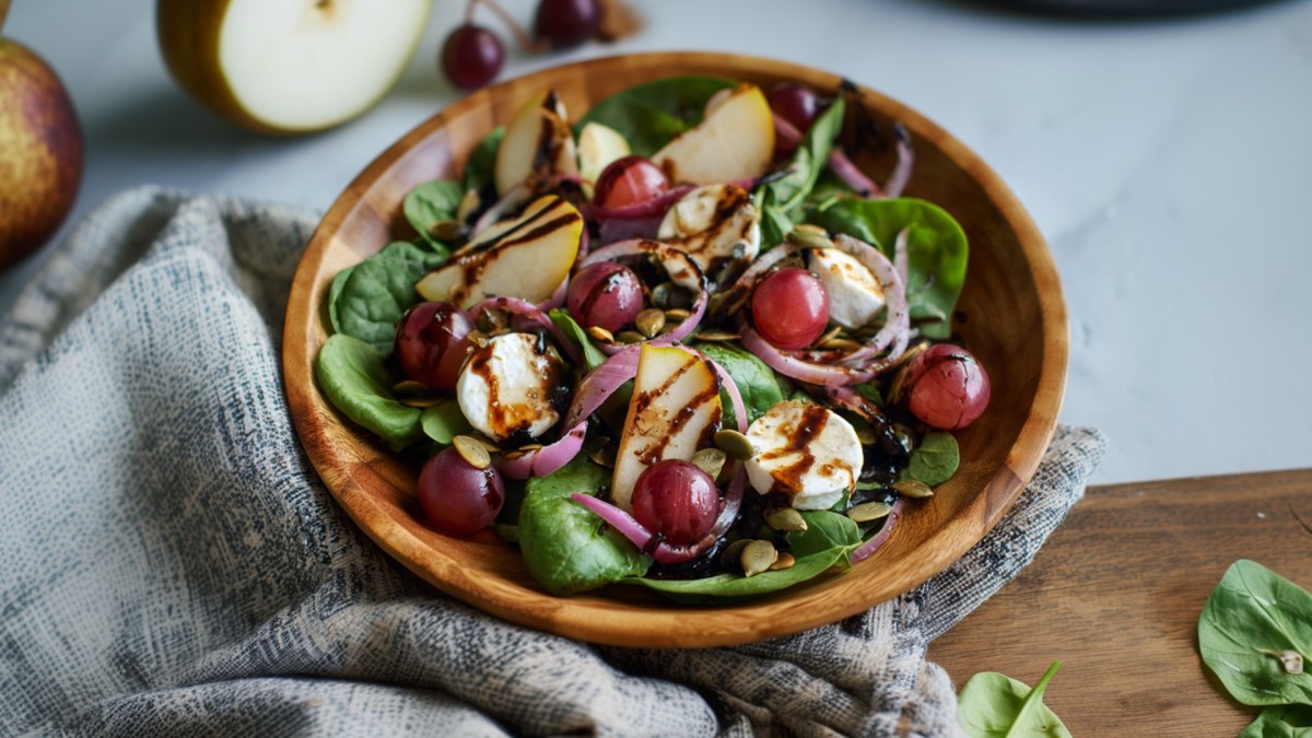 Eine Holzschüssel mit Herbstsalat mit Birne und Ziegenkäse. Im Hintergrund kann man eine Birne und einen Birnenschnitz erkennen sowie einige einzelne Trauben.