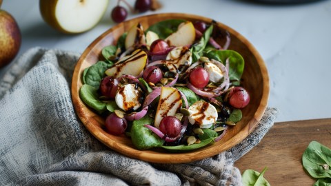 Eine Holzschüssel mit Herbstsalat mit Birne und Ziegenkäse. Im Hintergrund kann man eine Birne und einen Birnenschnitz erkennen sowie einige einzelne Trauben.