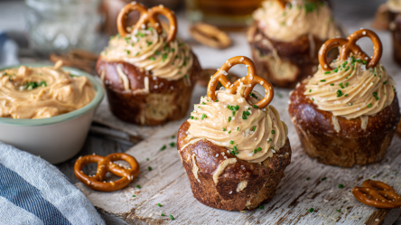 Vier herzhafte Brezel-Cupcakes auf eine hölzernen Unterlage. Links im Bild steht ein Schälchen mit Obazda. Davor liegt eine kleine Salzbrezel.
