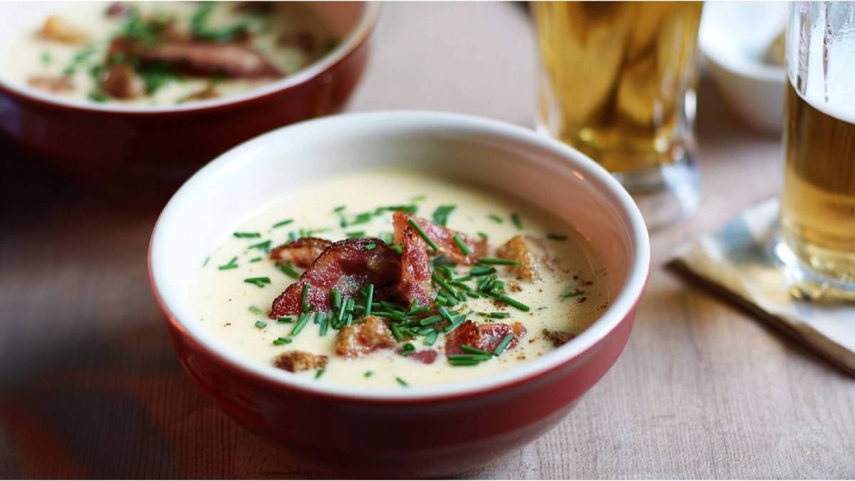 Schale mit Käse-Bier-Suppe mit Speck und Croûtons auf einem Tisch, Glas Bier daneben