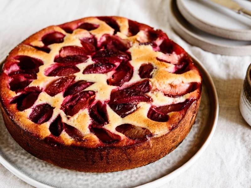 Ein ganzer Käsekuchen mit Pflaumen, der auf einem großen Teller platziert ist. Rechts daneben steht eine Schale mit Pflaumen. Im Hintergrund kann man Teller und Besteck erkennen.
