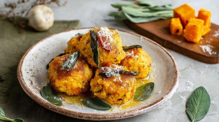 Ein Teller Kürbisknödel mit Salbeibutter. Im Hintergrund ist ein Holzbrett zu sehen, auf dem Kürbiswürfel und einige Blätter Salbei liegen. Außerdem kann man eine Knoblauchknolle erkennen.