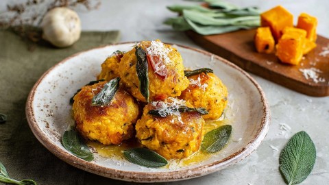 Ein Teller Kürbisknödel mit Salbeibutter. Im Hintergrund ist ein Holzbrett zu sehen, auf dem Kürbiswürfel und einige Blätter Salbei liegen. Außerdem kann man eine Knoblauchknolle erkennen.