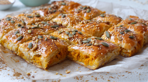 Ein, in Stücke geschnittener Kürbiskuchen vom Blech. Er ist mit gehackten Walnusskernen, Kürbiskernen und Sesam bestreut. Im Hintergrund sind zwei kleine Schalen zu sehen.