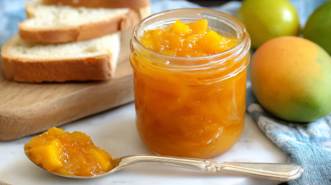Ein Glas Mango-Limetten-Marmelade mit einem Löffel mit der Marmelade daneben.
