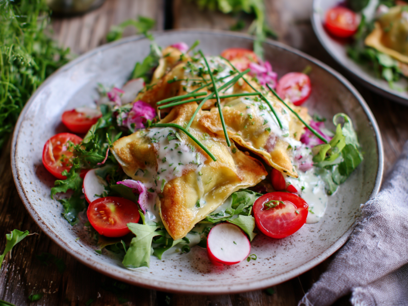 Maultaschensalat aus dem Airfryer auf einem grauen Teller. Drumherum liegen frische Kräuter verteilt. Im Hintergrund sieht man einen weiteren Teller mit dem Salat.