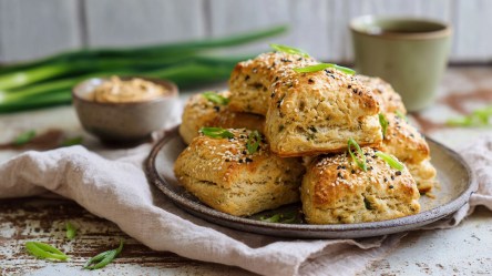 Ein Teller mit einem Stapel Miso-Scones, die mit Sesam und Frühlingszwiebeln garniert sind. Im Hintergrund kann man frische Frühlingszwiebeln, eine Schale mit heller Miso-Paste und einen Trinkbecher aus Keramik sehen.