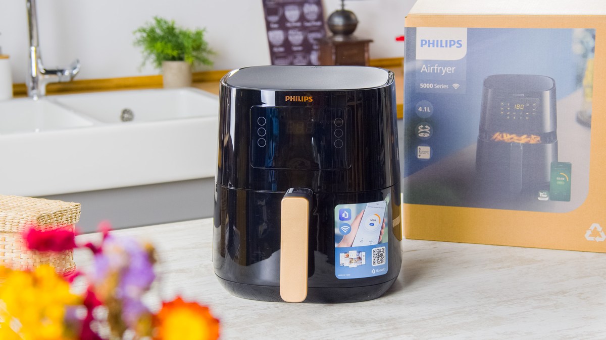 Ein Philips Airfryer 500 aus der Connected Series, der auf einer hellen Arbeitsplatte in einer Küche steht. Hinter dem Airfryer ist der Karton des Produkts platziert. Im Vordergrund kann man verschwommen einige bunte Blumen erkennen. Ansonsten kann man etwas vom Rest der Küche sehen.