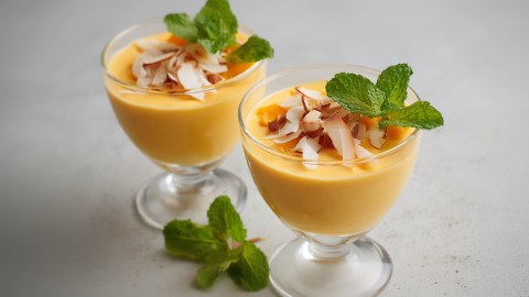 Zwei Dessertgläser mit Pfirsich-Mango-Mousse. Beide Portionen sind mit gerösteten Kokoschips und gerösteten Mandeln sowie frischer Minze garniert.