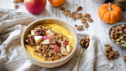 Eine Pumpkin-Pie-Joghurt-Bowl, die mit Apfelstücken, Granola und Walnüssen getoppt ist. Drum herum liegen ein Kürbis, ein Apfel, Nüsse und Granola verstreut.