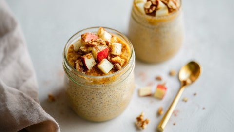 Ein kleines Glas mit Pumpkin Spice Chiapudding, der mit Apfelstückchen und Walnüssen getoppt ist. Nebenbei liegt ein Löffel. Im Hintergrund kann man ein weiters Glas mit Pumkin Spice Chiapudding sehen.