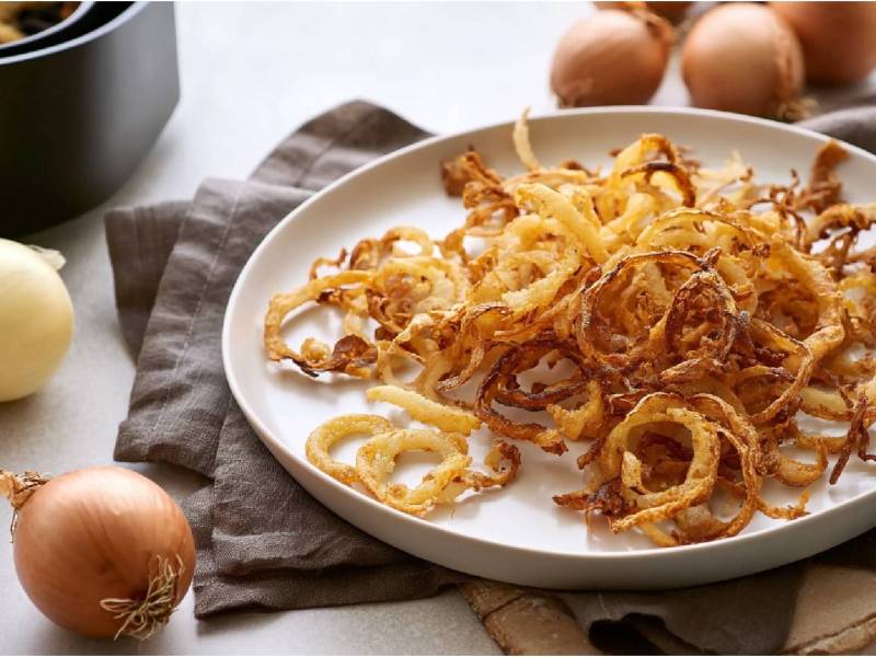 Schale mit Röstzwiebeln aus dem Airfryer, frische Zwiebeln und Airfryer im Hintergrund