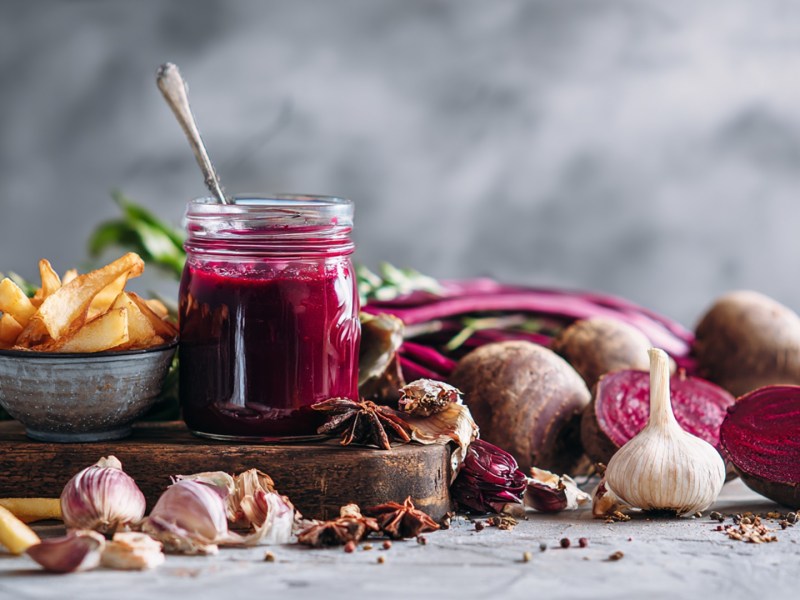 Ein Glas mit selbst gemachtem Rote-Bete-Ketchup, das auf einem Holzbrett steht. Daneben steht eine kleine Schale mit Pommes. Ringsum liegen Knoblauch, frische Rote Bete sowie Gewürze herum.