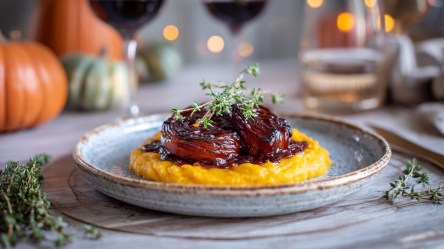 Ein Teller mit geschmorten Rotwein-Schalotten und Kürbispüree. Im Hintergrund kann man einige Dekokürbisse, Weingläser und eine Lichter erkennen.