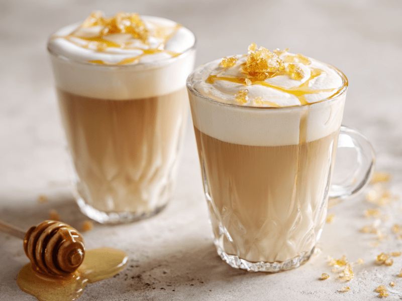 Zwei Gläser Salted Honey Latte auf einem hellen Untergrund. Auf dem Kaffee sind grobe Salzkörner zu sehen. Neben den Gläsern liegt ein Honiglöffel.