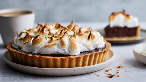 Eine Schoko-Baiser-Tarte, die auf einem Teller platziert ist. Im Hintergrund kann man ein Stück Tarte auf einem Teller sowie eine Tasse Kaffee sehen.