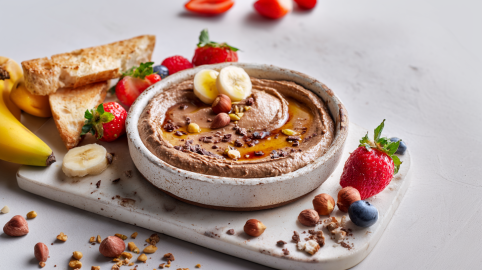 Eine Schale Schoko-Hummus auf einem weißen Brett. Der Hummus ist mit Bananenscheiben und Nüssen angerichtet. Um die Schale herum liegen Erdbeeren, Bananen, Blaubeeren sowie Toaststücke.