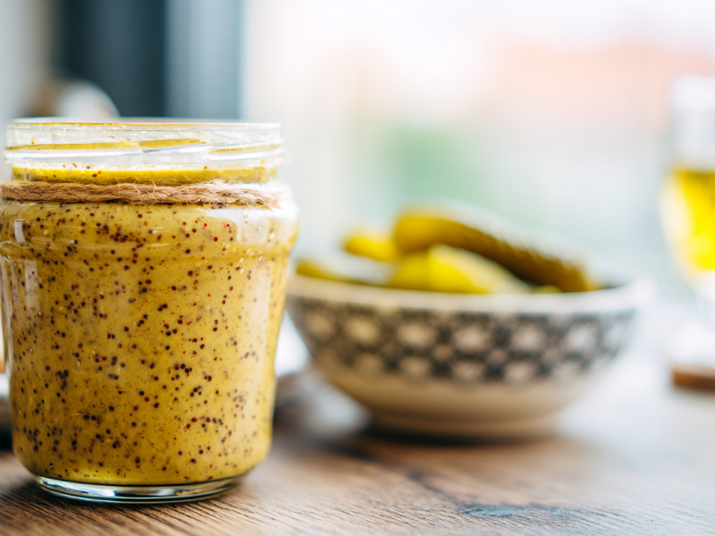 Ein Glas mit Senf aus Gurkenwasser. Im Hintergrund ist ein Schälchen mit Gewürzgurken und ein Bier zu sehen.