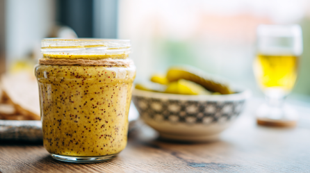 Ein Glas mit Senf aus Gurkenwasser. Im Hintergrund ist ein Schälchen mit Gewürzgurken und ein Bier zu sehen.