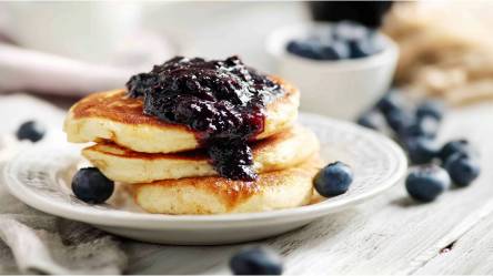 Teller mit Skyr-Pancakes mit Blåbärssylt und Blaubeeren in einer Schale dahinter