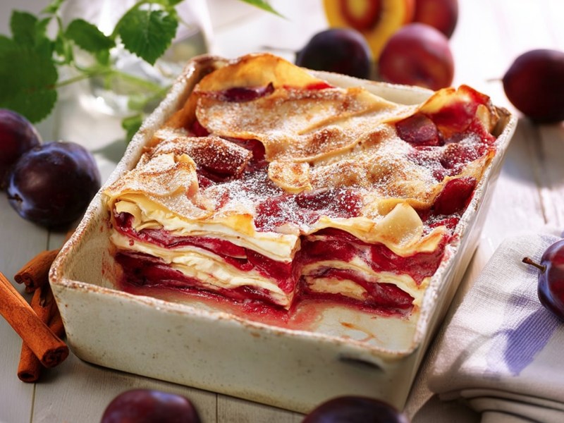 Eine Auflaufform mit einer süßen Pflaumen-Lasagne. Ringsum liegen frische Pflaumen, Zimtstangen und Minze verteilt.