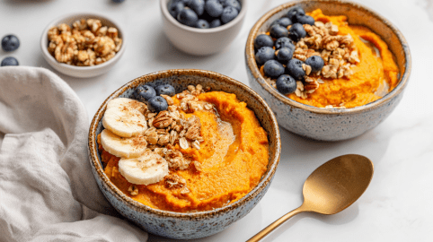 Zwei Schüsseln mit Süßkartoffel-Frühstücks-Bowls. Außen herum liegt ein Löffel, eine Stoffserviette und eine Schüssel mit frischen Heidelbeeren.