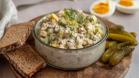 Schale mit Thunfisch-Eiersalat auf einem Tisch, Scheiben Brot, Gewürzgurken und ein aufgeschnittenes ei drumherum