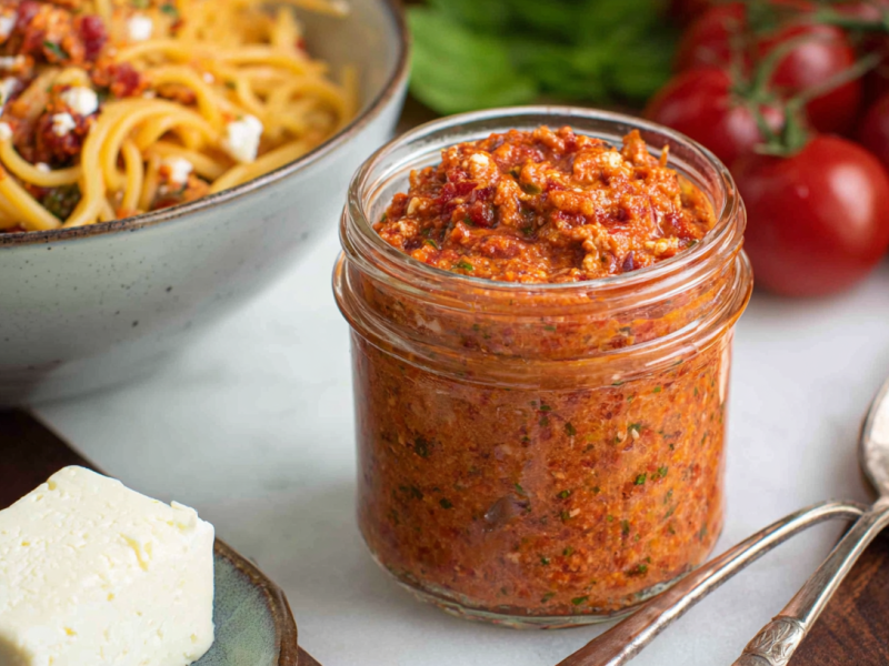 Ein Glas Tomaten-Feta-Pesto vor einer Schüssel Nudeln im Hintergrund.