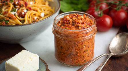 Ein Glas Tomaten-Feta-Pesto vor einer Schüssel Nudeln im Hintergrund.