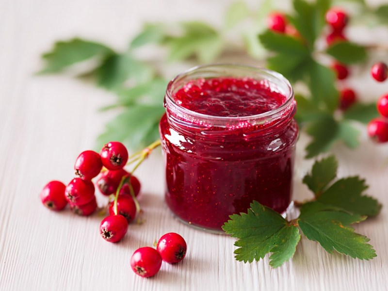 Ein Glas mit Weißdorn-Marmelade. Ringsum liegen ein paar Weißdornbeeren verteilt.