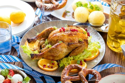 Ein Wiesnhendl auf einem Teller. Es ist umgeben von Orangenscheiben, Granatapfelkernen, Bier, Knödeln und Brezeln.