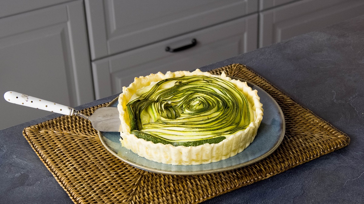 Eine Zucchini-Ricotta-Tarte auf einem grauen Teller auf einer Küchenmatte. Die Tarte ist angeschnitten und ein Kuchenheber liegt bereit, sie zu servieren.