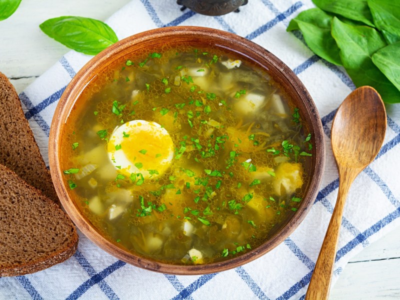 Eine Schale andalusische Hühnersuppe mit Fleisch, Eiern und Nudeln. Daneben liegt ein Löffel und zwei Scheiben Brot.