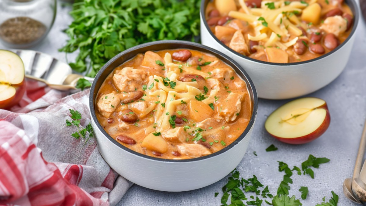 Eine Schüssel Apfel-Hähnchen-Chili, das mit frischen Kräuter garniert ist. Ringsum liegen frische gehackte Kräuter, aufgeschnittene Äpfel, ein Küchenhandtuch. Eine zweite Schüssel Chili steht im Hintergrund.