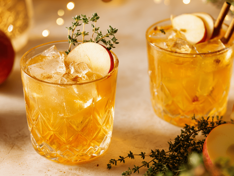 Zwei Gläser mit Apfel-Zimt-Cocktail mit Gin. Darum liegen frische Äpfel und ein Thymianzweig als Dekoration.